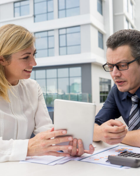 Smiling woman showing male colleague tablet screen in cafe outdoors. Businesspeople working and sitting at table with building in background. Business people working outdoors concept.
